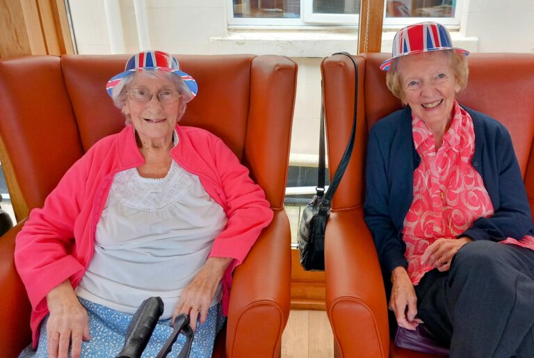 Two elderly women sit in orange chairs at the care home, both wearing Union Jack-themed hats and smiling as they enjoy a relaxed afternoon, dressed in casual attire.