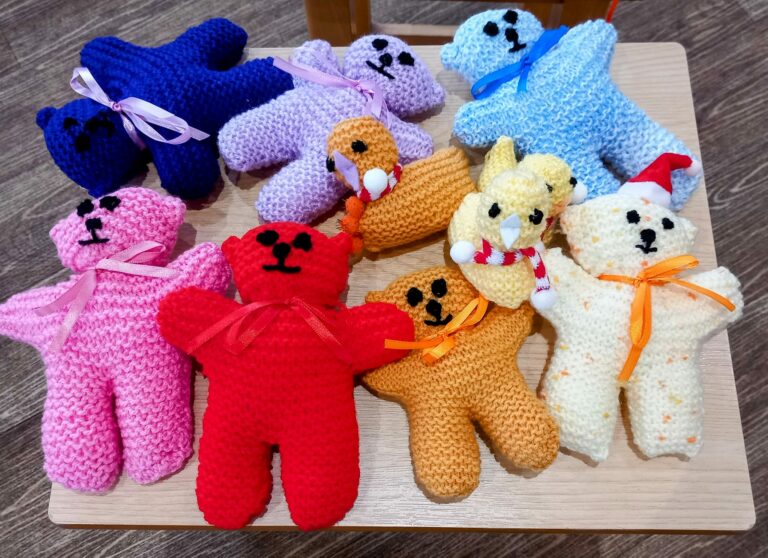 A variety of colourful knitted teddy bears with ribbons, some wearing hats, are arranged on a wooden surface at the Abbeyfield assisted care home.