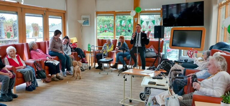 A group of elderly people are seated in a room at an Abbeyfield assisted care home, with a man speaking at the front. Party decorations adorn the walls, and a dog stands amongst the group.