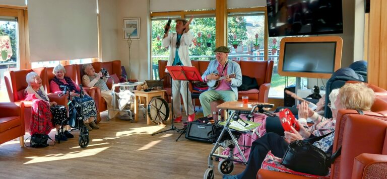 In a sunlit room at the Abbeyfield residential care home, a group of elderly residents enjoy the soothing respite of live music. One performer sings and plays guitar while another, seated, complements the melody with an instrument.