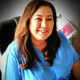 A person with long dark hair smiles warmly, wearing a blue shirt, seated in an office setting that exudes the welcoming atmosphere of Abbeyfield House.