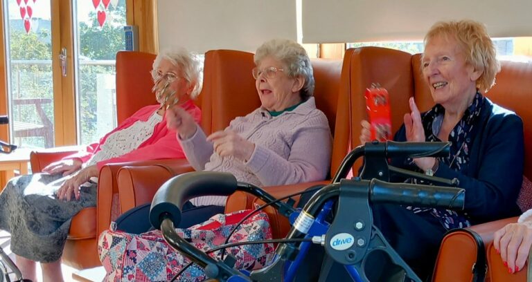 Three elderly women sitting in armchairs, smiling and holding shakers, enjoying their time in a cosy care home, with a walking frame nearby.