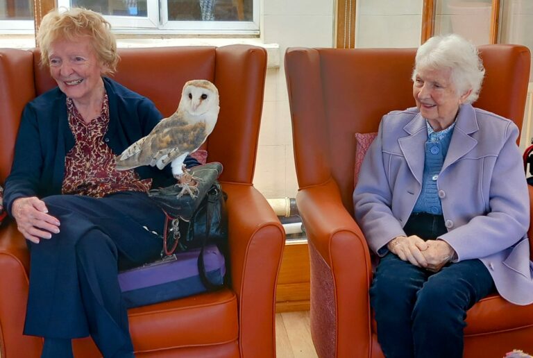 Two elderly women sit in armchairs at the care home, one of them with a barn owl perched gently on her lap.