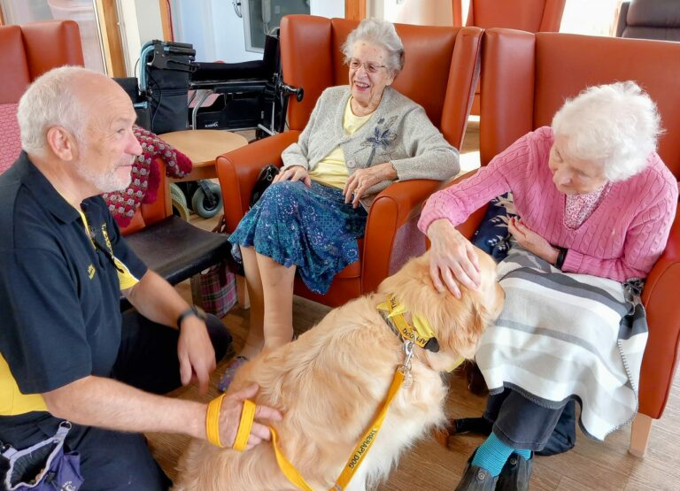 A man with a golden retriever visits two elderly women in a residential care home. One woman strokes the dog while the other smiles warmly. A wheelchair is visible in the background, adding to the sense of comfort and companionship that fills the room.