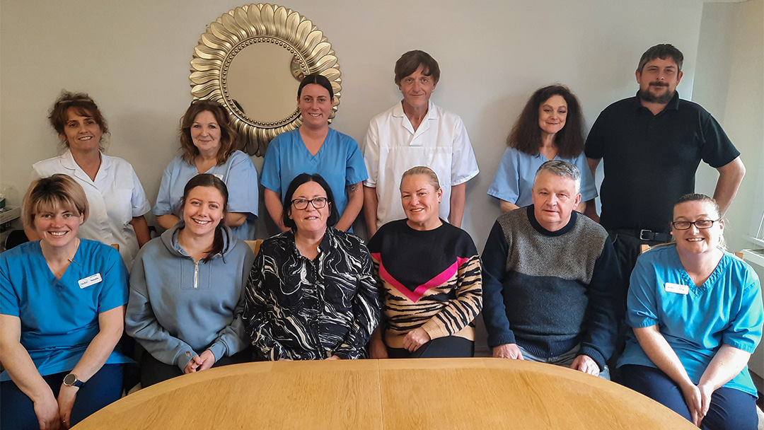 A group of twelve people, some in blue uniforms, pose around a table in the care home. A decorative mirror is on the wall behind them.