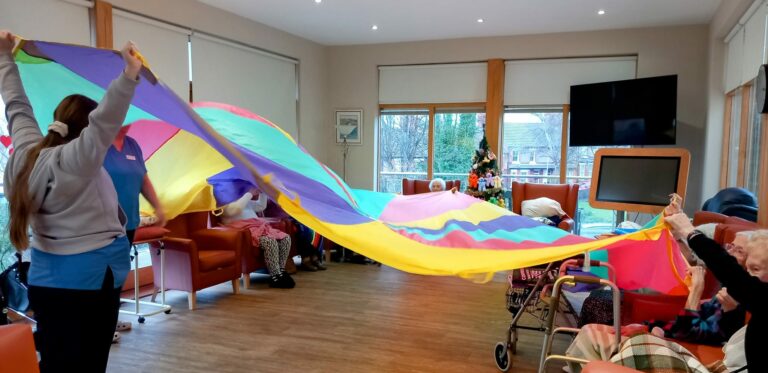 People in a room at the residential care home hold a colourful parachute cloth. The setting includes armchairs, a telly, and large windows with trees visible outside.