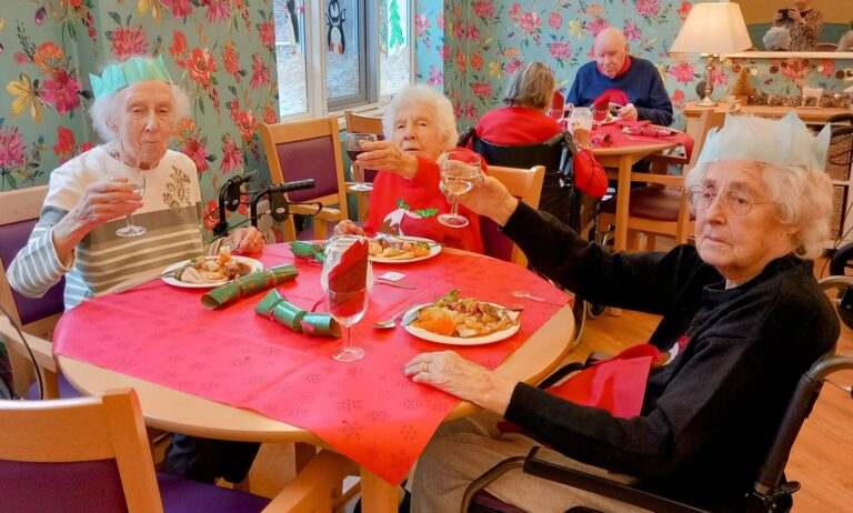 Elderly women in a Wirral care home sit at a festive table, raising glasses amidst holiday decorations. Plates of food fill the scene, and fellow residents add to the cheerful background.