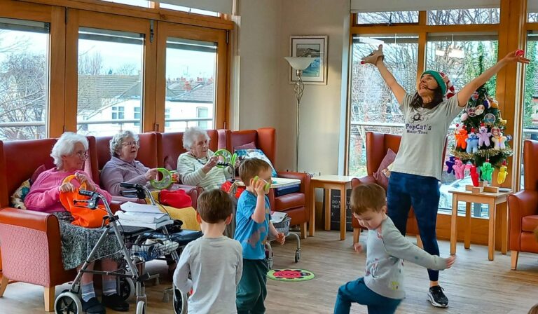 An instructor leads a lively dance activity with children at a Wirral care home, while elderly women seated nearby join in using shakers, all set in a bright room with large windows and a beautifully decorated tree.