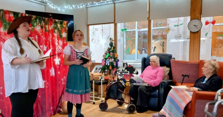 Two people in fancy dress perform before an audience in a room decorated with Christmas motifs at the Abbeyfield residential care home. Two elderly women sit watching, one in a pink jumper and the other wrapped in a colourful blanket, enjoying the festive entertainment.