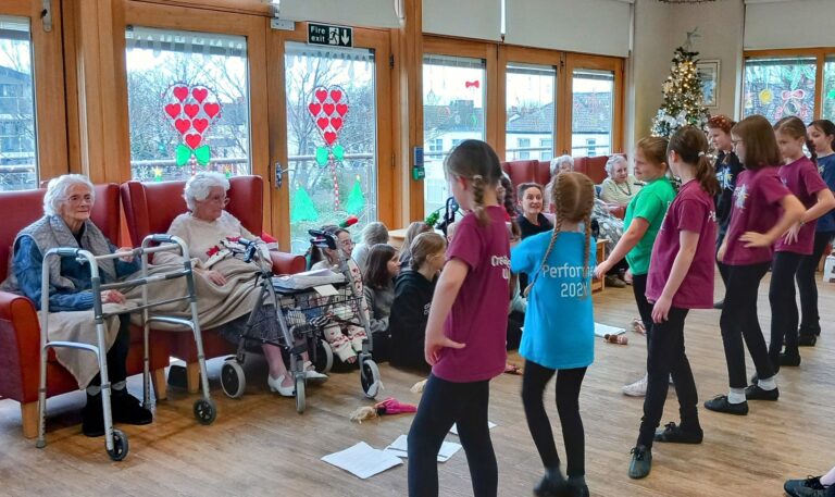 Children perform in front of elderly residents at the Abbeyfield residential care home, a room adorned with festive decorations and heart-shaped window stickers.