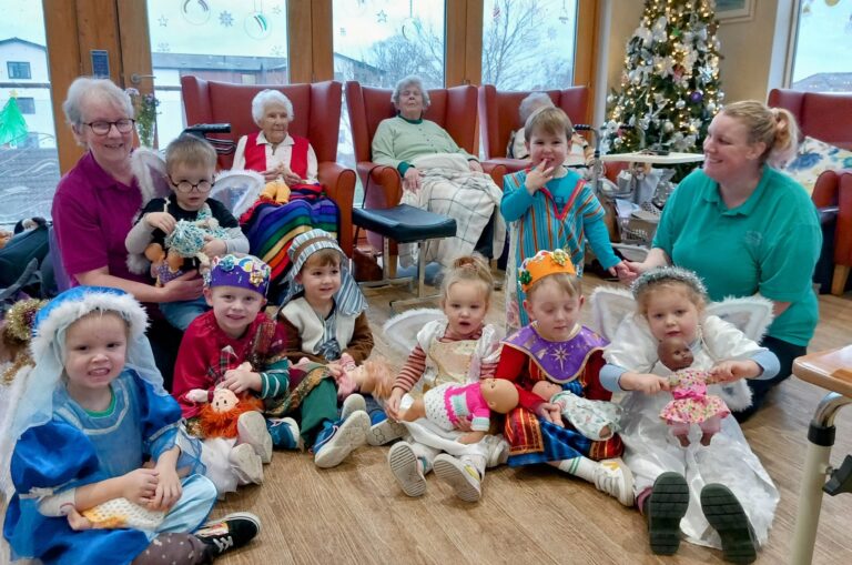 Children in fancy dress sit with dolls and adults near a glittering Christmas tree at the Abbeyfield Wirral care home. Elderly women are seated in the background, while two adults join the children on the floor, adding to the festive warmth of this special gathering.