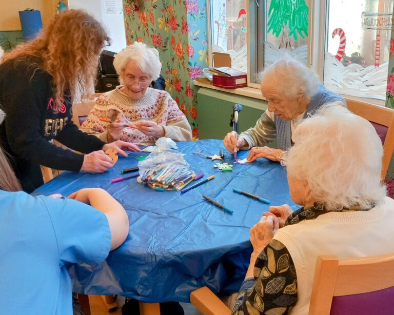 Elderly women sit around a table with a blue cloth, engaging in crafts with marker pens at an Abbeyfield residential care home. A woman assists them while holiday decorations add a festive touch to the scene.