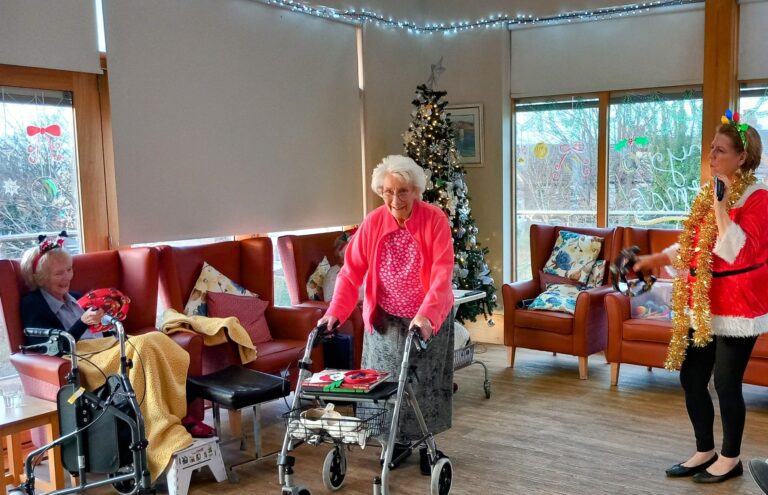 In a cosy room at the Wirral care home, an elderly woman with a walking frame smiles, joined by another in festive attire. Nearby, someone sits in a wheelchair, wrapped in a blanket. The Christmas tree twinkles warmly, embodying the holiday spirit shared at Abbeyfields respite care gathering.