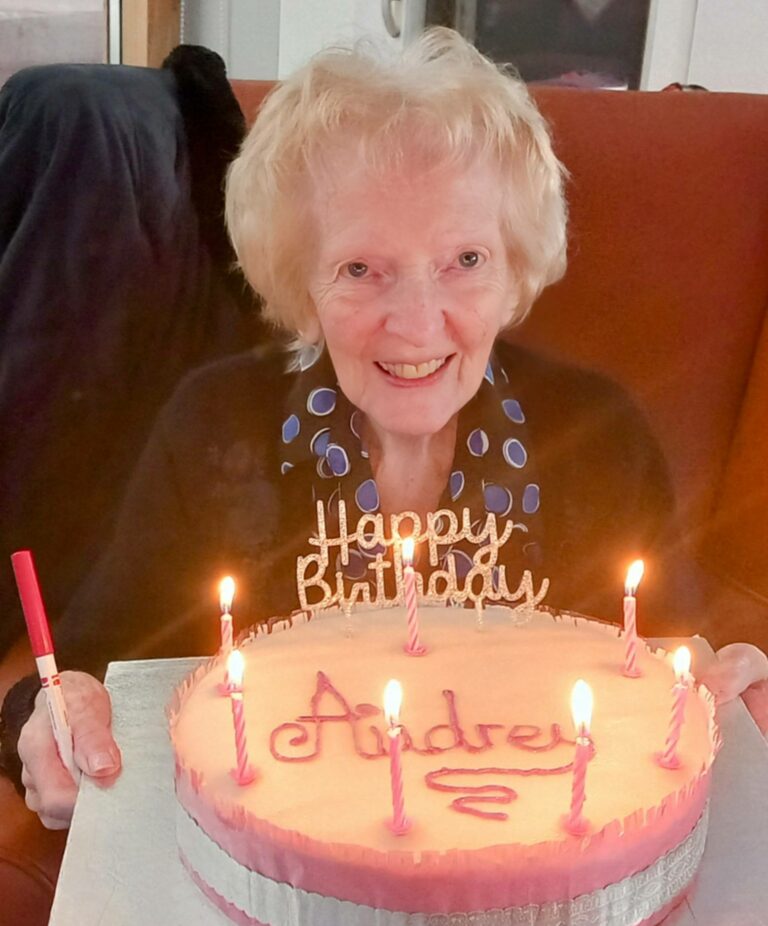 Elderly woman smiles warmly whilst holding a birthday cake with lit candles and Happy Birthday Audrey decoration, celebrating another cherished year at the Abbeyfield residential care home.