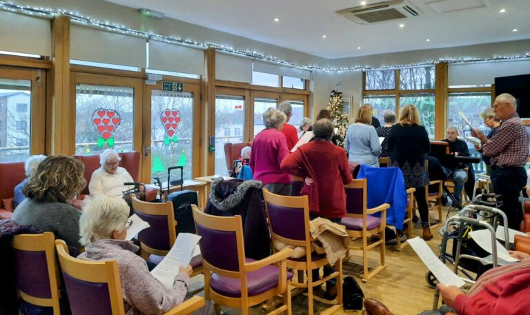 A group of elderly people gather in a cosy residential care home, standing or sitting with papers, surrounded by holiday lights and window decals. In the background, a calming melody flows as someone plays the piano.