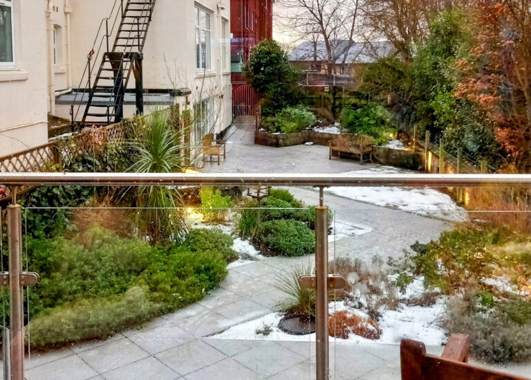 A garden with snow-dusted pathways, surrounded by greenery and trees, reminiscent of a peaceful Wirral care home. Metal railings in the foreground, buildings on the left, and a fire escape complete the serene scene beneath an overcast sky.