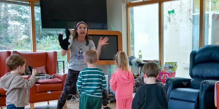 In a cosy living room adorned with holiday decorations, a woman animatedly entertains four children with a puppet show. The children watch her closely, captivated. This heartwarming scene unfolds in an Abbeyfield care home, where joy and community come together during respite care moments.