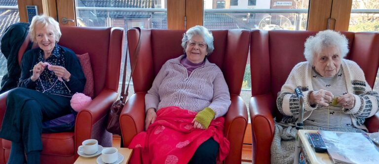 Three elderly women sit in armchairs, knitting and smiling, with cups of tea on a nearby table in the well-lit room of a Wirral care home.