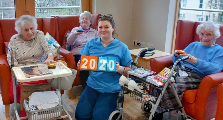 In an Abbeyfield residential care home, a carer proudly holds number blocks arranged as 2020. Three elderly women are seated around the room, engaged with various activity items on their tables and chairs, enjoying the lively atmosphere.