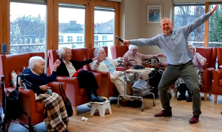 A man energetically poses with a microphone in front of smiling elderly people seated in armchairs at the Abbeyfield care home, their laughter resonating through the room filled with large windows.