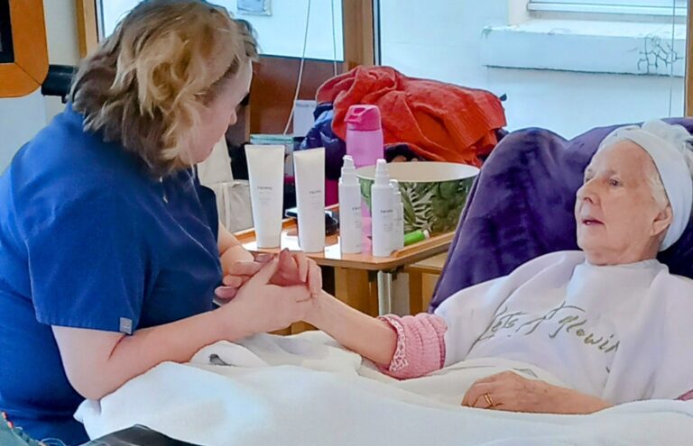 A carer in blue scrubs holds the hand of an elderly woman wearing a headband, seated comfortably under a blanket. In the Wirral care home, various skincare products rest on the table beside them, offering comfort and tranquillity during this respite care moment.