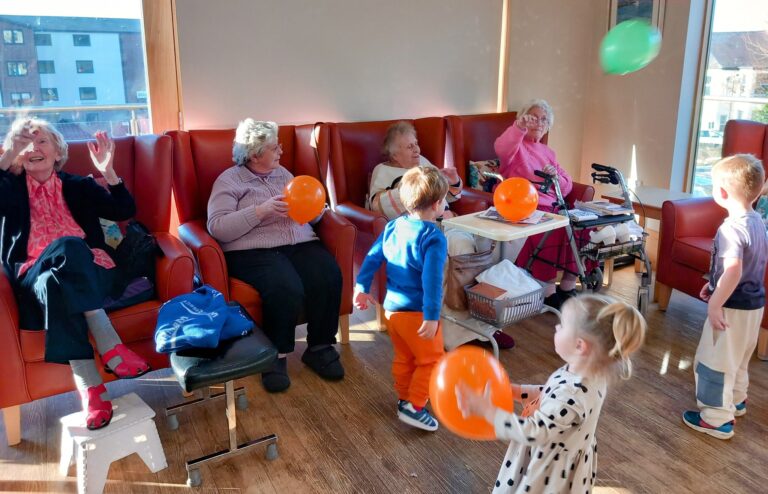 Elderly women sit in chairs, watching young children play with balloons in the bright room of the Abbeyfield care home.