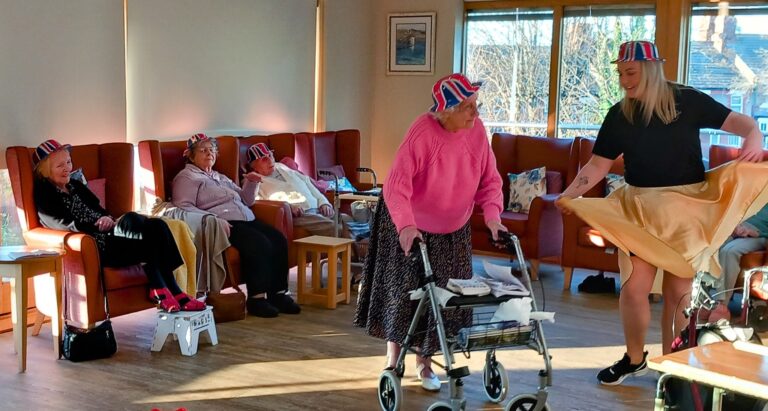 In a vibrant room with large windows at a Wirral care home, elderly women, some adorned with Union Jack hats, joyfully watch and participate in a lively dance session led by a young woman.