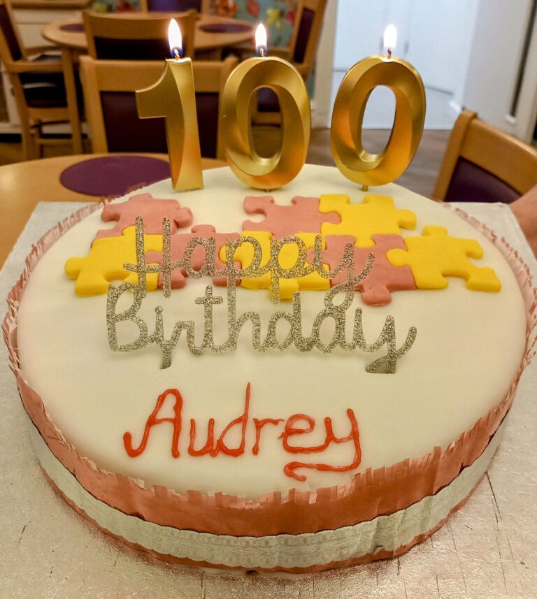 Birthday cake with white icing, decorated with Happy Birthday Audrey and puzzle pieces. Topped with candles shaped as 100, lit and standing proudly on a table at the care home, where everyone gathers to celebrate this remarkable milestone.