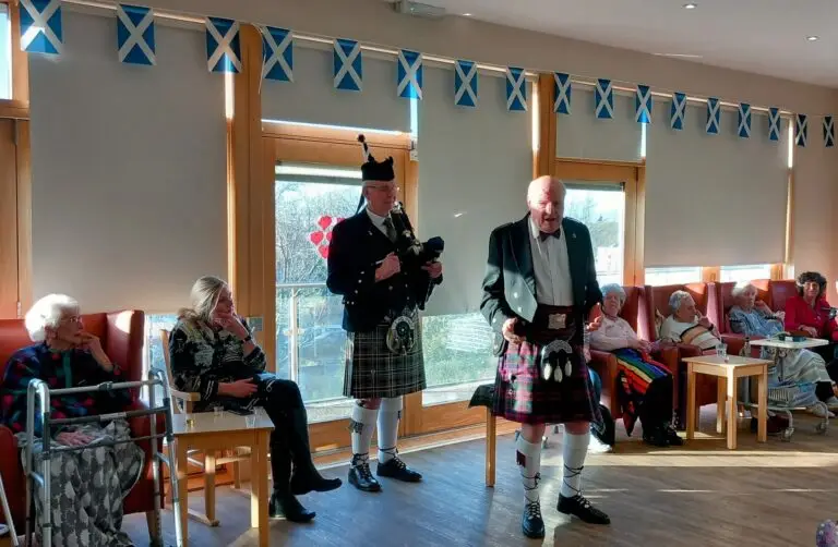 A group of people gather in an Abbeyfield room with a Scottish theme. A man in a kilt performs near the centre while another, also in traditional attire, stands with a microphone. Flags hang from the ceiling, creating a vibrant atmosphere at this assisted care home event.