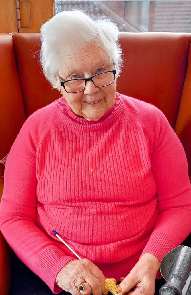 An elderly woman with white hair and glasses is sitting on a red bench, wearing a pink jumper, and knitting with yellow wool.