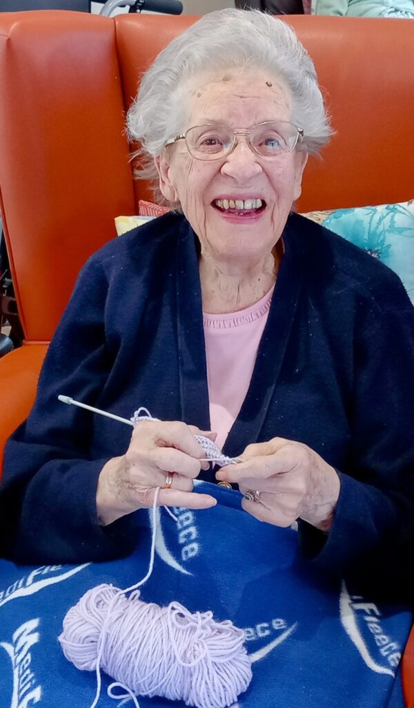 Elderly woman sitting in an orange chair, smiling whilst crocheting with light purple yarn.