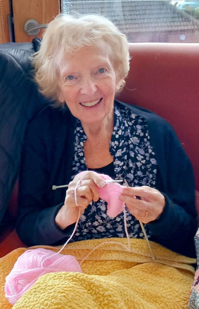 An elderly woman knitting with pink wool, sitting on a red sofa, and smiling at the camera.