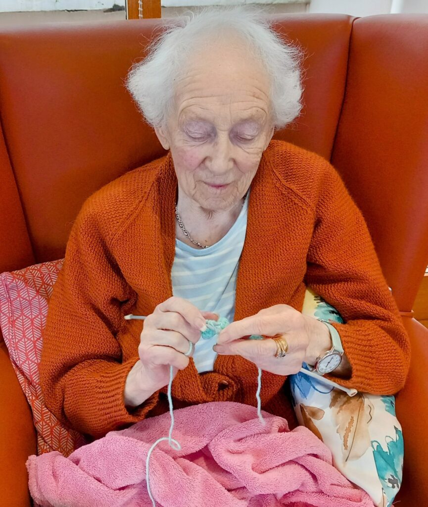 Elderly woman with white hair sitting in a brown chair, wearing an orange cardigan, concentrating on knitting with light-coloured yarn.