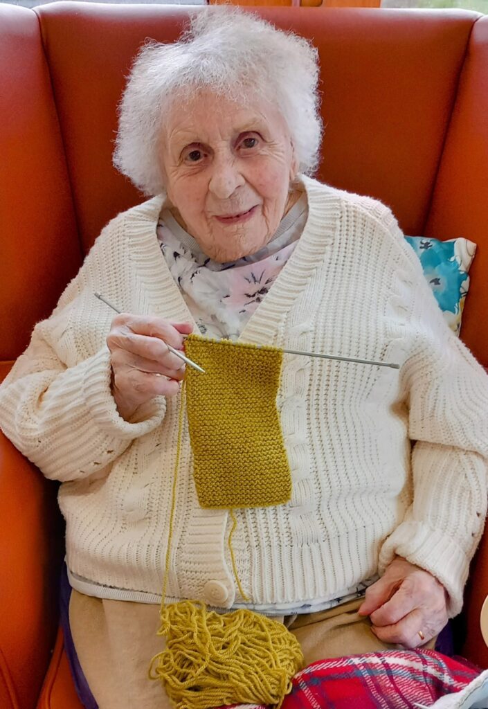 An elderly woman with white hair sits on an orange chair, knitting a yellow piece of fabric. She is wearing a white jumper.