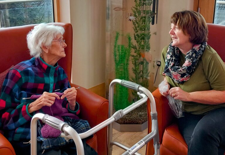 An elderly woman knits in a red armchair at the Wirral Care Home, chatting with a visitor holding a plastic carrier bag. Nearby, a walking frame and an aquarium contribute to the cosy atmosphere.