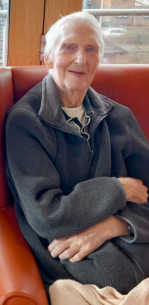 Elderly person with grey hair, wearing a dark grey jumper, sitting on a brown chair in a room with large windows.