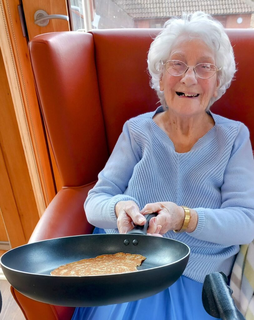 An elderly woman sitting on a red chair at Abbeyfields Lear House smiles warmly, her light blue jumper contrasting with the frying pan holding a perfectly cooked pancake. Celebrating Pancake Day with joy in her heart and spectacles perched on her nose, she embodies the spirit of tradition.