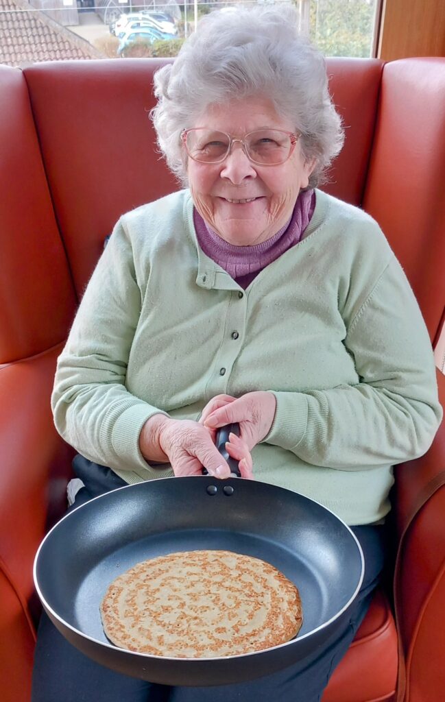 An elderly woman wearing glasses and a green jumper sits in a red chair at Abbeyfield Lear House, holding a frying pan with a cooked pancake, celebrating Shrove Tuesday.