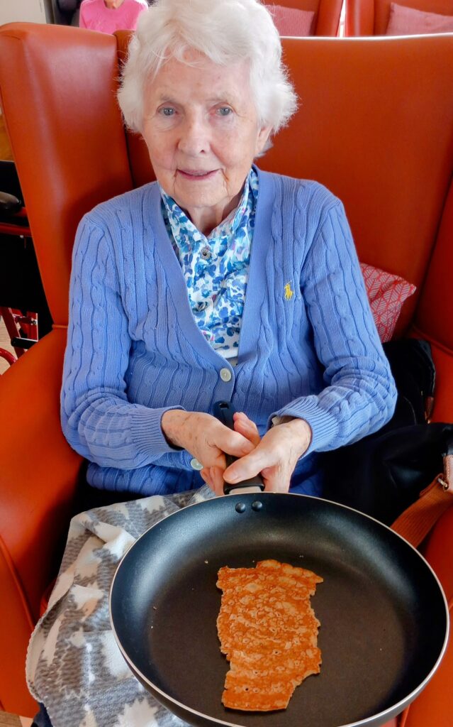 An elderly woman seated in a chair at Abbeyfield Lear House, cheerfully holding a frying pan with a cooked pancake inside, celebrating Shrove Tuesday.