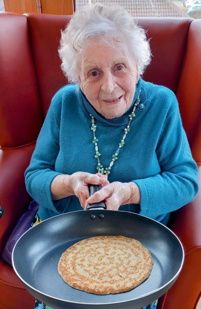 An elderly woman beams while sitting on a red chair at Lear House, holding a frying pan with a perfectly cooked pancake. Dressed in a blue jumper and beaded necklace, she embraces the joy of Pancake Day.