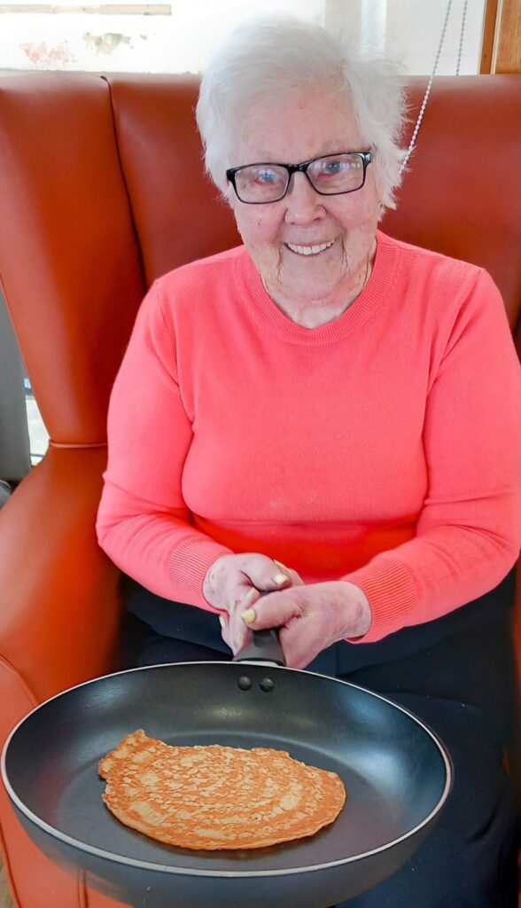 An elderly person wearing a coral jumper and glasses smiles while holding a frying pan with a pancake on it, celebrating Pancake Day at Abbeyfield Lear House, sitting comfortably on a brown chair.