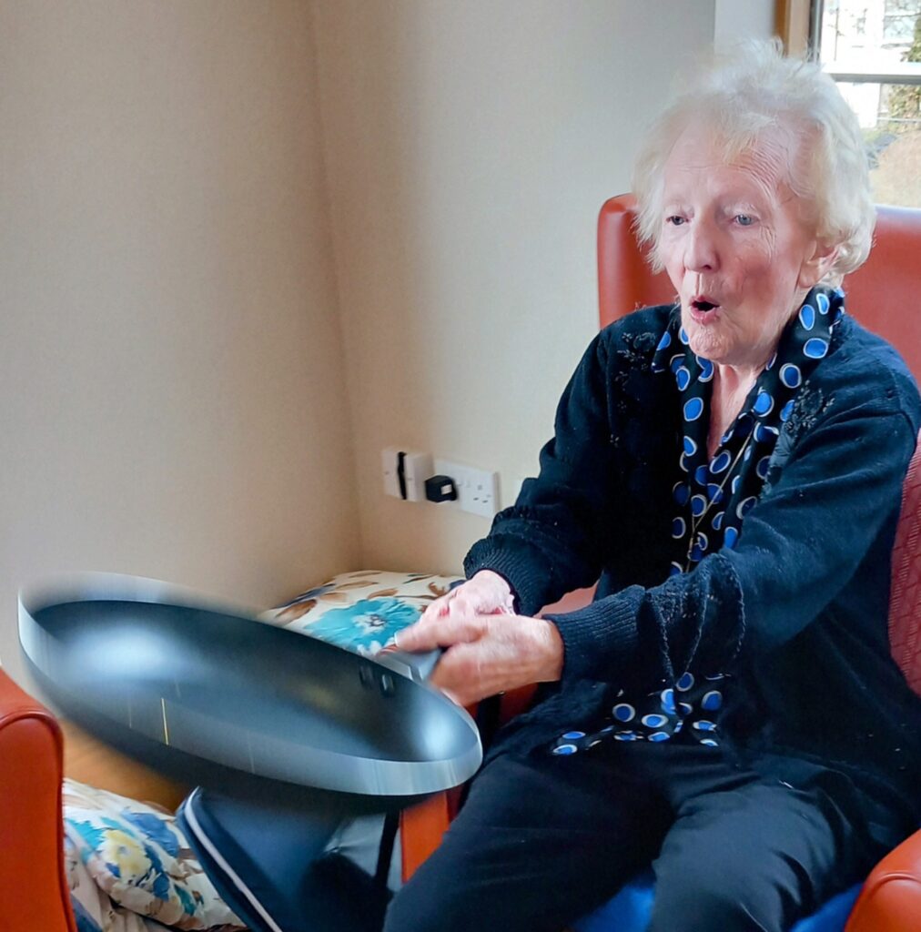 Elderly woman sitting in a chair at Lear House, playfully holding a black frying pan with both hands, an expression of surprise or excitement lighting up her face, as if shes ready to flip pancakes for Shrove Tuesday.