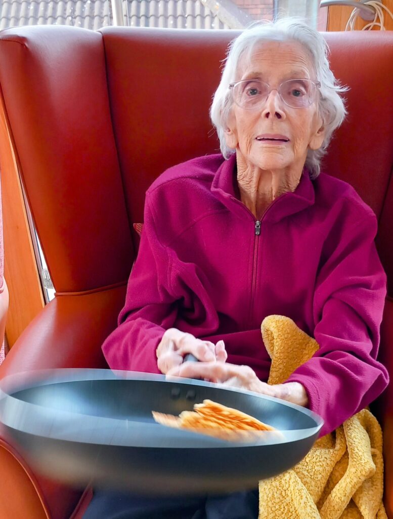 An elderly person in a burgundy jumper sits in a red chair at Lear House, deftly holding a pan with a pancake mid-flip, celebrating Shrove Tuesday.