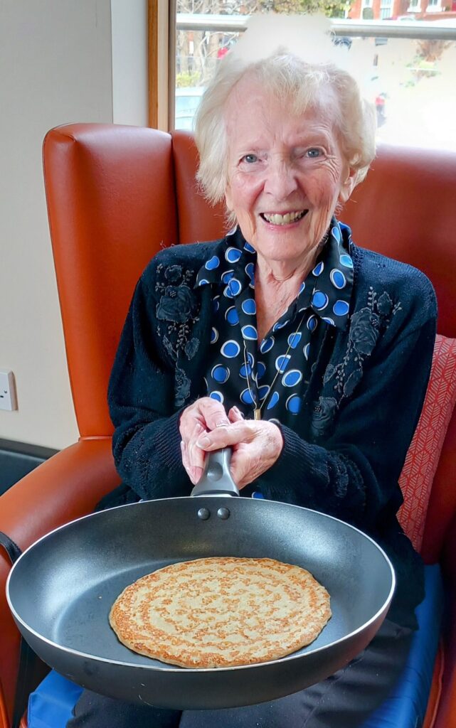 Elderly woman smiling whilst holding a frying pan with a cooked pancake, celebrating Pancake Day on an orange chair at Lear House.