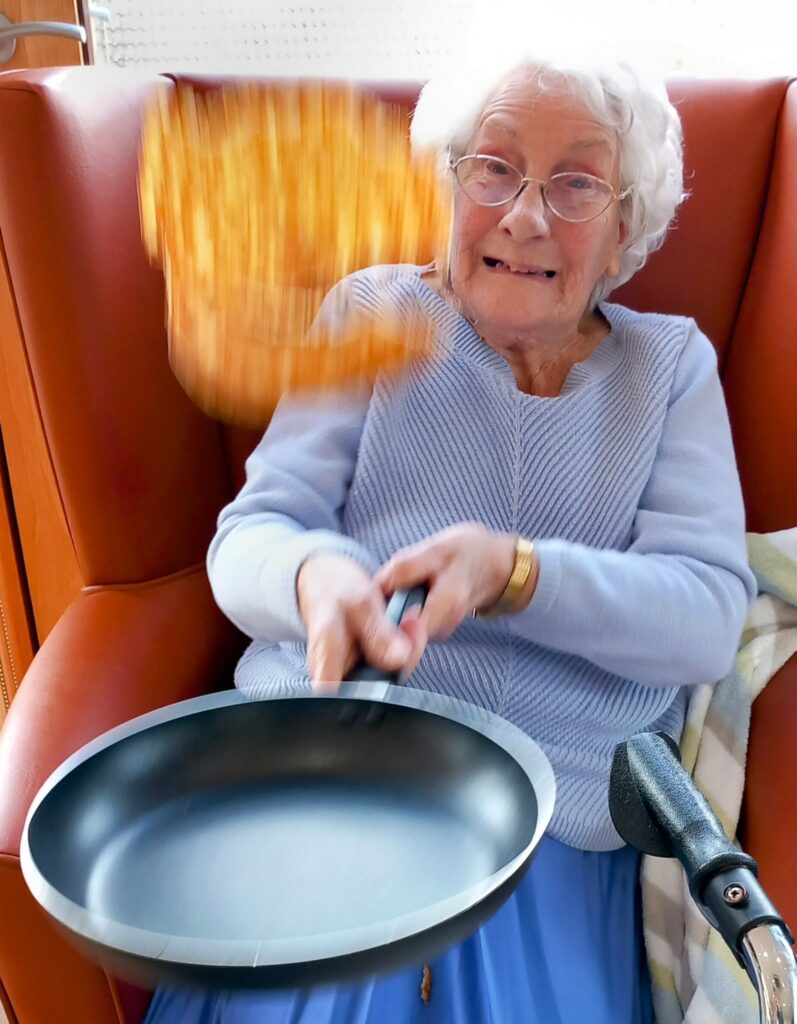 An elderly woman sitting in a chair at Abbeyfield Lear House, flipping a pancake in a frying pan on Shrove Tuesday.