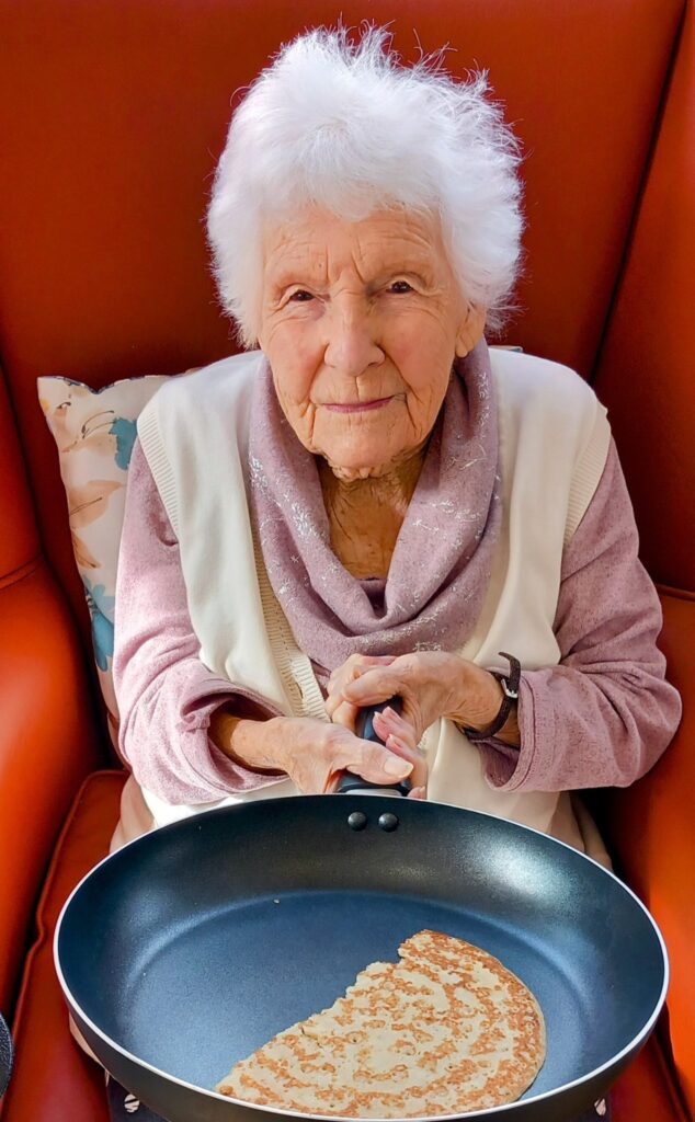 An elderly woman sits comfortably in a chair at Abbeyfield Lear House, holding a frying pan with a partially cooked pancake inside, celebrating Shrove Tuesday with joy and culinary skill.