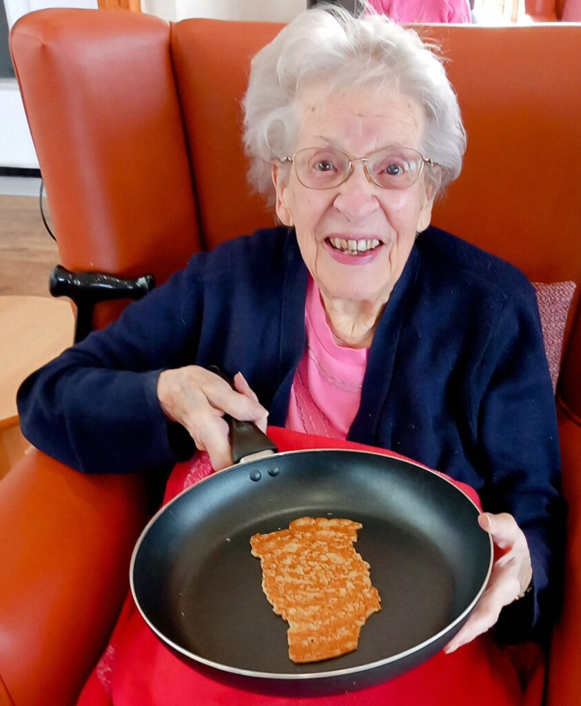 An elderly woman at Abbeyfield Lear House sits in a chair, holding a frying pan displaying a pancake shaped like the state of Illinois. Its Pancake Day, and her creative breakfast masterpiece brings smiles all around.