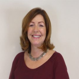 A smiling woman with shoulder-length brown hair, wearing a silver necklace and a dark red top, stands against a plain background, her warmth and kindness reflecting the welcoming atmosphere of an Abbeyfield residential care home.