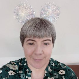 A person with short grey hair, radiant in a floral top and shiny tinsel accessories resembling antennae, stands against a plain backdrop. Their vibrant style brings joy to the Abbeyfield care home, where respite care allows them moments of creative expression.